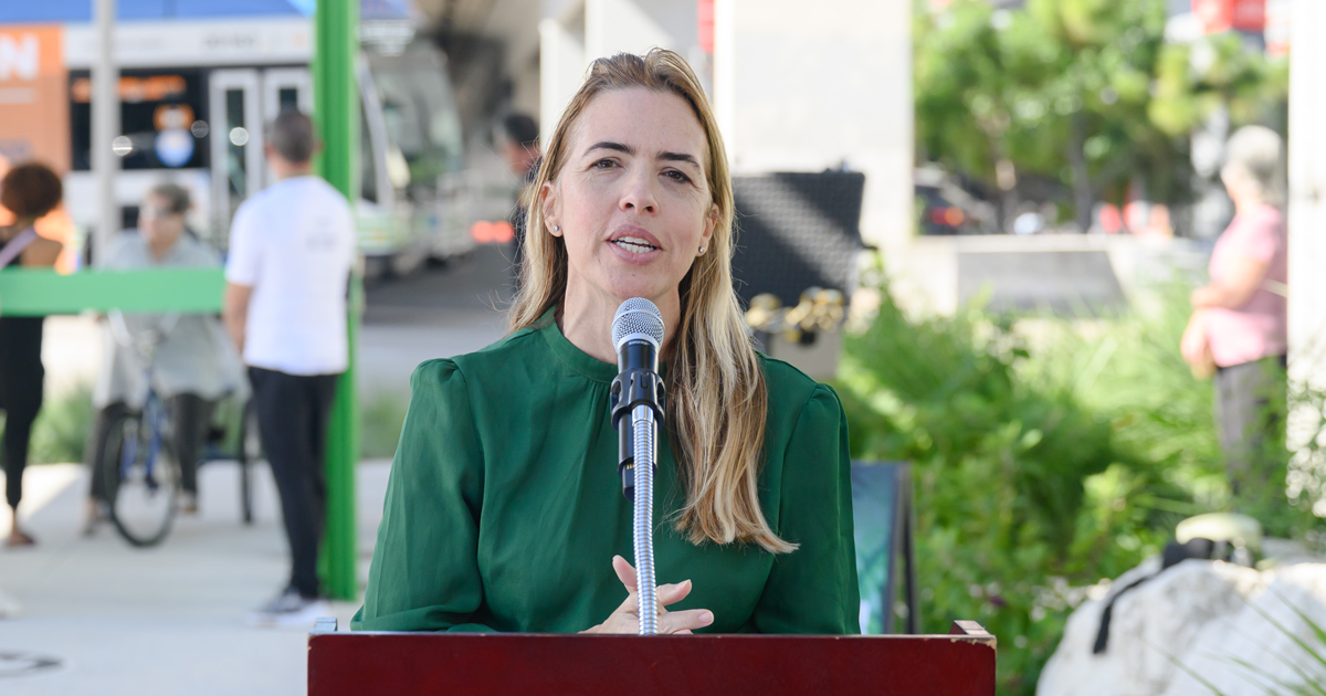 Miami-Dade County Commissioner Raquel Regalado speaks into a microphone at an outdoor podium during a community event. She wears a green blouse, with greenery, event equipment, and a sidewalk visible in the background.
