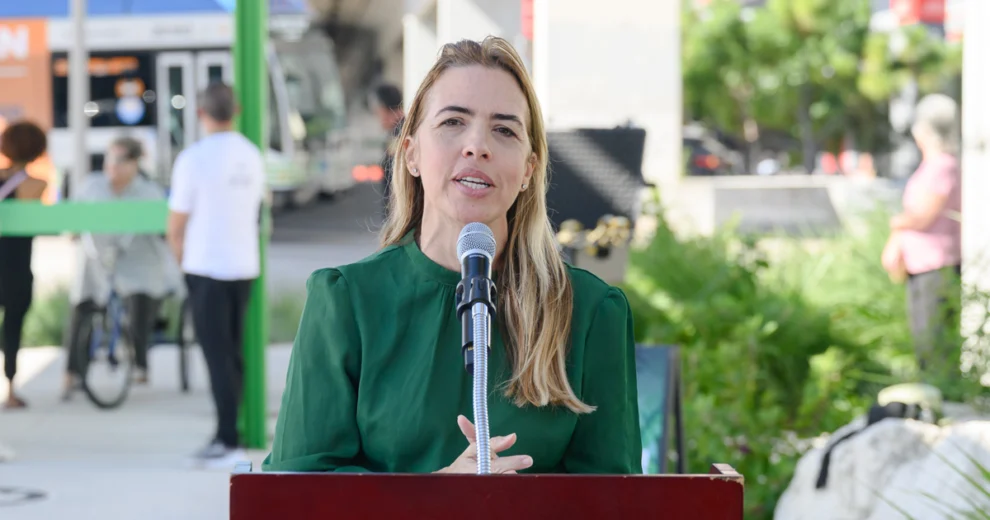 La comisionada del condado de Miami-Dade, Raquel Regalado, habla ante un micrófono en un podio al aire libre durante un evento comunitario. Lleva una blusa verde; de ​​fondo, se observan vegetación, equipamiento del evento y una acera.