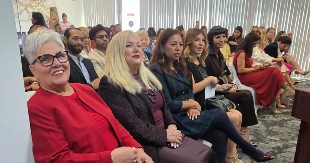 Attendees sit in a conference room during a presentation, with several women seated in the front row and a large audience behind them.
