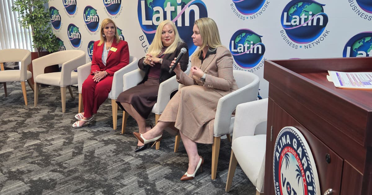 Three women sit in chairs during a panel discussion in front of a Latin Business Network backdrop, with one woman holding a microphone.