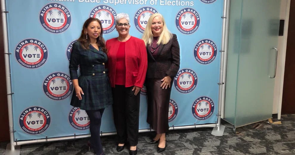 Three women stand together in front of a backdrop displaying Alina Garcia, Miami-Dade County Supervisor of Elections, and “VOTE” logos.