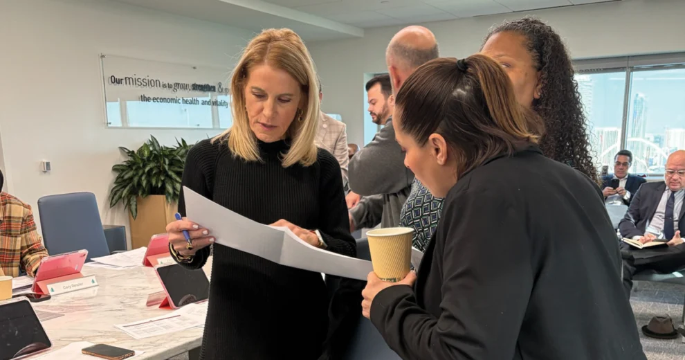 Miami-Dade County Commissioner Vicki Lopez reviews a document with a colleague during a meeting in a conference room as other attendees sit around the table.
