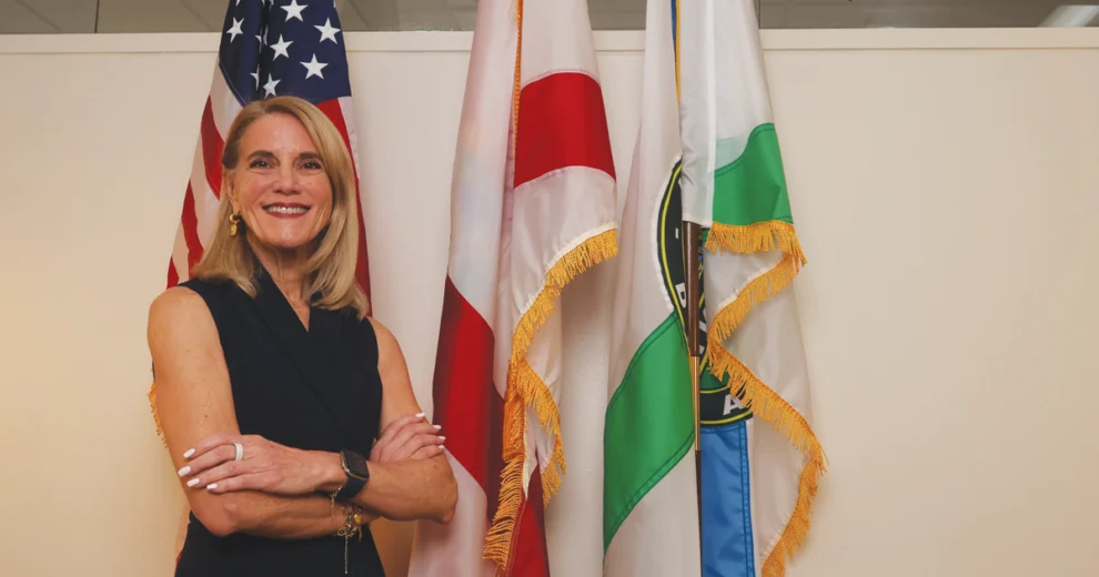 Miami-Dade County Commissioner Vicki Lopez smiles with her arms crossed while standing in front of the American flag, Florida state flag, and Miami-Dade County flag.