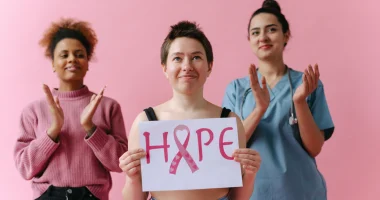 Three women stand against a pink background; the woman in front holds a sign reading &ldquo;HOPE&rdquo; with a pink breast cancer awareness ribbon, while the two women behind her applaud.