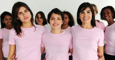 A diverse group of women wearing pink shirts stand together and smile at the camera, symbolizing breast health and breast cancer awareness.