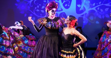 Una colorida actuación cultural hispana en el Adrienne Arsht Center, con bailarines que lucen elaborados trajes y maquillaje inspirado en el Día de los Muertos sobre un escenario iluminado de azul.