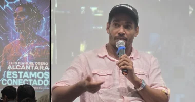 Luis Manuel Otero Alcántara habla por un micrófono dirigiéndose al público durante un coloquio sobre el documental "Luis Manuel Otero Alcántara: Estamos Conectados". Viste una camisa rosa claro y una gorra negra, y gesticula mientras se proyecta una imagen promocional de la película en una pantalla a su lado.