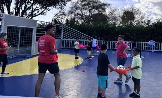 Un grupo de niños participa en una actividad grupal en una cancha al aire libre, utilizando una tela roja para transportar juntos un balón de fútbol, ​​mientras otros observan y juegan en las cercanías. Un entrenador o un joven mayor, vestido con una camiseta roja, permanece de pie guiando la actividad; al fondo, se aprecian conos y una portería.
