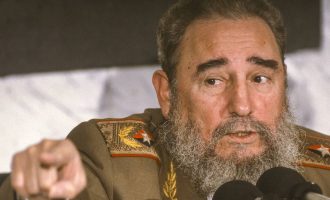 Fidel Castro in a military uniform gestures forward with his hand while speaking into microphones. His expression appears intent and engaged, as if emphasizing a statement during a speech or press conference.