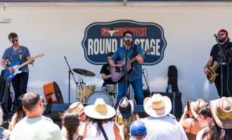 Una banda de música country actúa en vivo en un escenario al aire libre rotulado como «Round Up Stage», con músicos tocando guitarras y batería.