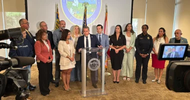 Miami-Dade County Tax Collector Dariel Fernandez speaks at a podium during a press conference announcing a humanitarian relief effort for earthquake victims in Venezuela. He is joined by local officials, community leaders, first responders, and nonprofit representatives standing behind him.