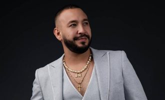 Un hombre con una barba pulcramente recortada posa sobre un fondo oscuro, vistiendo un traje gris claro con una camisa de escote en V pronunciado.