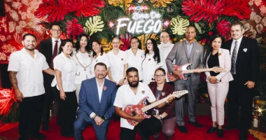 A group of Seminole Hard Rock Hollywood team members and executives pose for a group photo in front of a colorful tropical display featuring a neon &ldquo;Zona de Fuego&rdquo; sign. Several team members hold custom-designed electric guitars decorated with vibrant artwork.