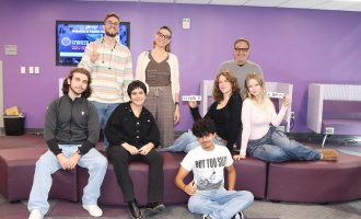 Un grupo de estudiantes y un miembro del profesorado posan juntos dentro de un salón del campus con paredes moradas.