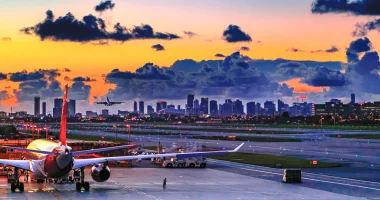An airplane sits on the runway at Miami International Airport at sunset, with airport lights and the Miami skyline visible in the background beneath colorful clouds.