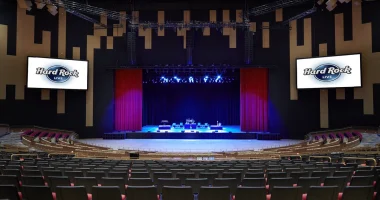 Vista interior panorámica del Hard Rock Live en el Seminole Hard Rock Hotel & Casino Hollywood, que muestra el teatro vacío con filas de asientos orientadas hacia un escenario intensamente iluminado y enmarcado por cortinas rojas.