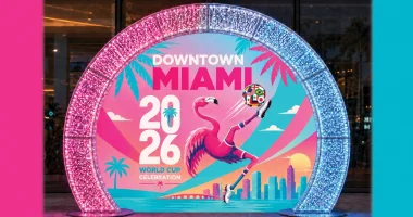 Una vibrante instalación titulada "Celebración de la Copa Mundial 2026 en el centro de Miami" presenta un flamenco rosa que equilibra un colorido balón de fútbol, ​​con un telón de fondo de palmeras, vistas al océano y el horizonte urbano de Miami. Unos arcos iluminados en rosa y azul enmarcan la instalación, creando un escenario festivo ideal para tomar fotografías y celebrar la Copa Mundial de la FIFA 2026.