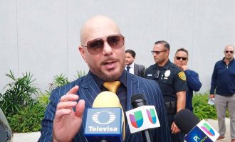 Un hombre calvo con gafas de sol y traje sostiene con confianza dos micrófonos, listo para hablar o actuar.