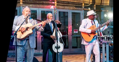 Musicians perform live at Rhythm & Roots, with three guitarists and singers playing in front of a building entrance.