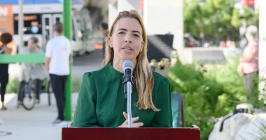 Miami-Dade County Commissioner Raquel Regalado speaks into a microphone at an outdoor podium during a community event. She wears a green blouse, with greenery, event equipment, and a sidewalk visible in the background.