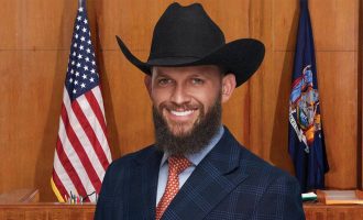 Robert Kelbert wearing a black cowboy hat and suit smiles in a courtroom setting with U.S. and state flags behind him.