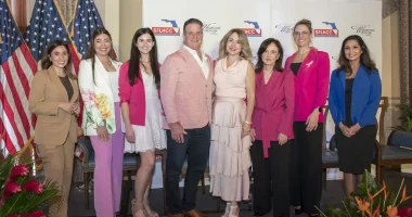 Eight individuals pose for a group photo at an event hosted by the South Florida Hispanic Chamber of Commerce and Hispanic Women&rsquo;s Society. The group stands before a branded backdrop with American flags on the left and floral arrangements in the foreground.