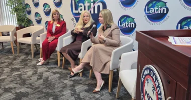 Three women sit in chairs during a panel discussion in front of a Latin Business Network backdrop, with one woman holding a microphone.