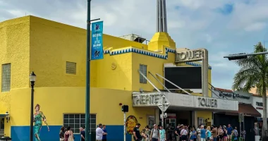 Vista exterior diurna del histórico "Tower Theater", en la Calle Ocho de Little Havana, Miami. El edificio, de color amarillo brillante y estilo Art Déco, presenta detalles ornamentales geométricos en azul y blanco, el destacado letrero luminoso con el nombre TOWER THEATER y una multitud de personas reunidas en el exterior.