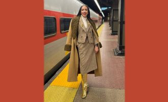 Yli Liriano de pie en una estación de metro, con un abrigo largo y botas doradas.