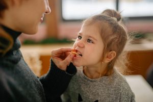 A young girl playfully bites into a slice of an orange that her mom feeds her.
