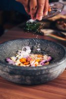 A person sprinkles salt into a mortar and pestle with onions, spices, and herbs, to grind and mix.
